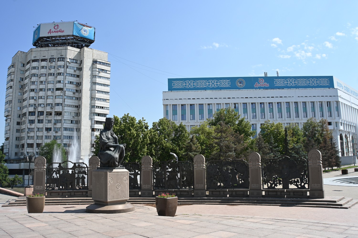 Širokoplošné mestské námestie so sochou sedacej ženy v černom, čiernymi kovanými plotmi, stromami, modernou budovou s modrým znakom Almaty v pozadí a vysokou obytnou vežou vľavo.