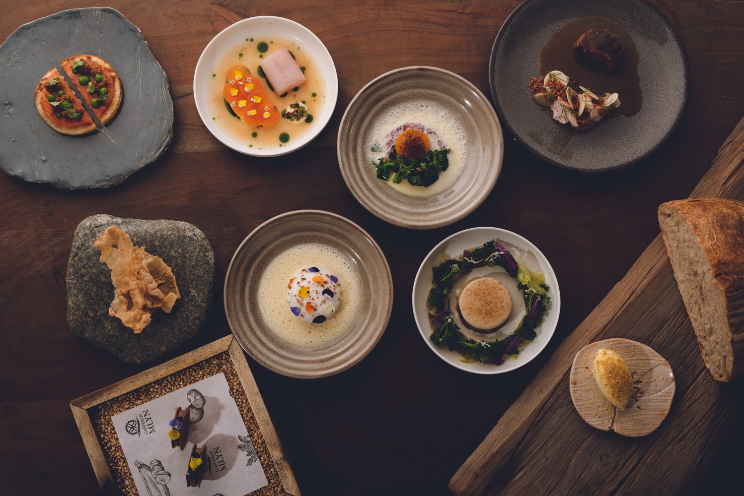 Sedem artísticky upravených chodov na rôznych keramických tanieroch a miskách rozložených na drevenom stole, vedľa chleba a kamennej dekorácie.