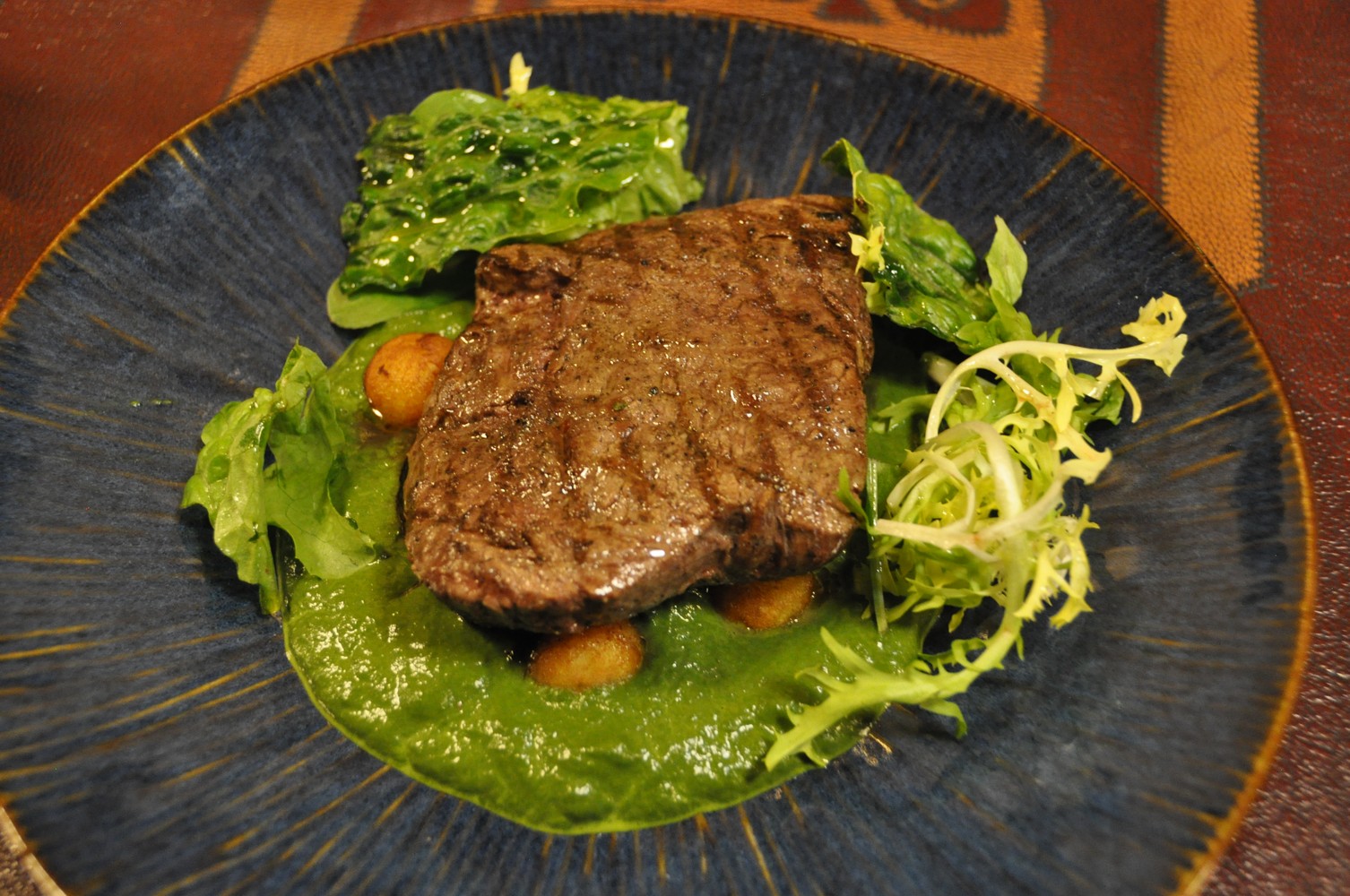 Seared meat steak on dark blue plate with vibrant green herb sauce, edible flowers, microgreens, and roasted potatoes, warm-toned fabric backdrop.