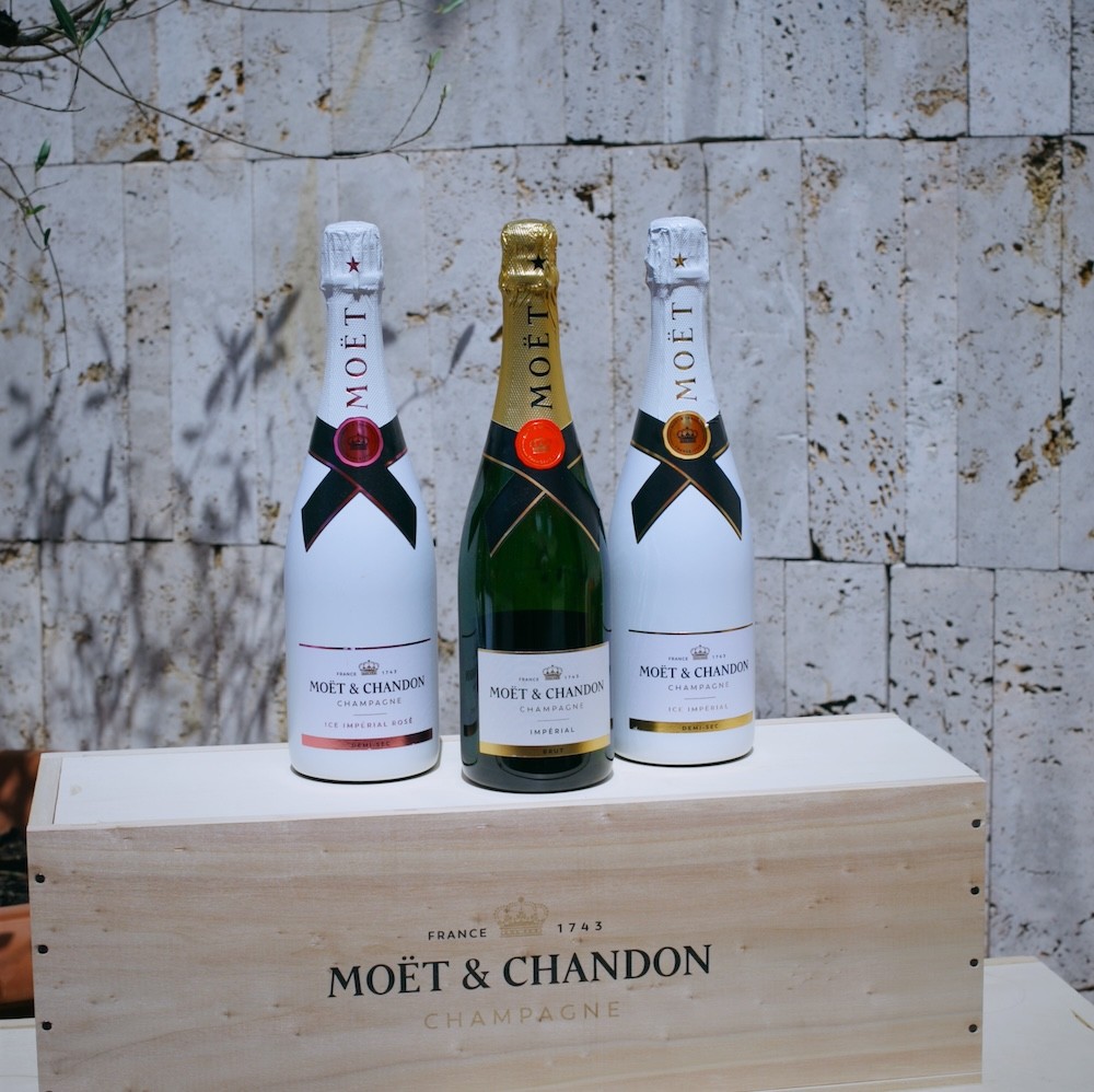 Tri fľaše šampanského Moët & Chandon v bielej a zelené flaši stojace na drevenom boxe s označením "MOËT & CHANDON CHAMPAGNE" pred textúrovanou betónovou stenou.