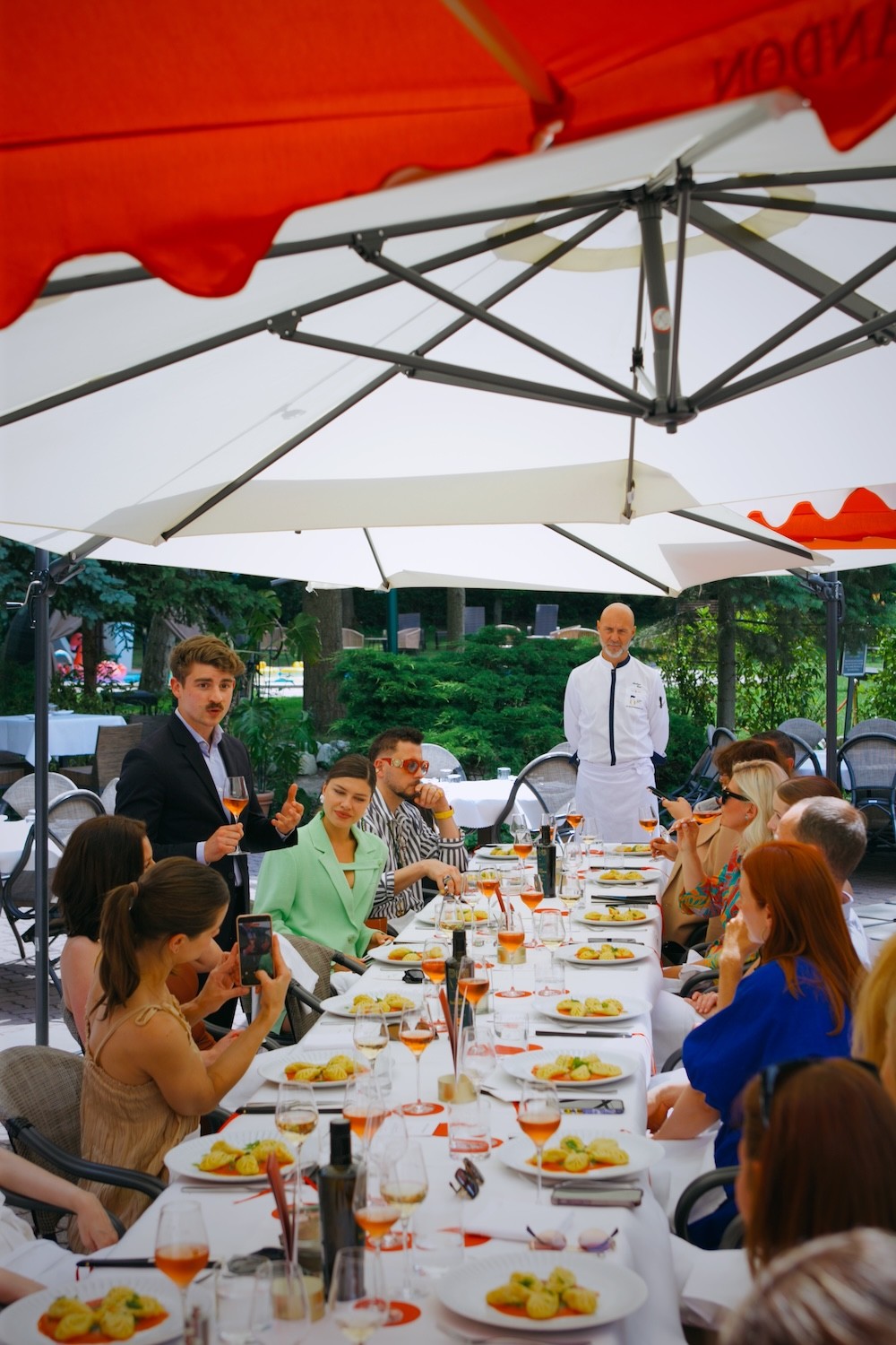 Outdoor dining scene under red and white umbrellas with guests seated at a long table with white tablecloth, plates of food, and glasses of rosé wine.